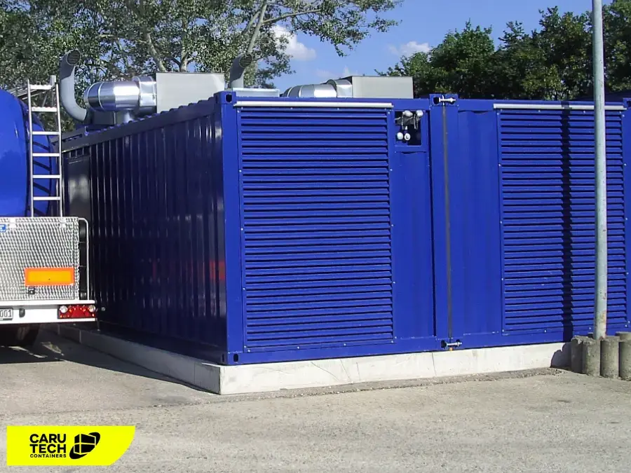 Blauer Notstromaggregat-Container mit großen Lüftungsgittern und Abgasrohren, neben einem Tanklastwagen auf einem Industriehof.