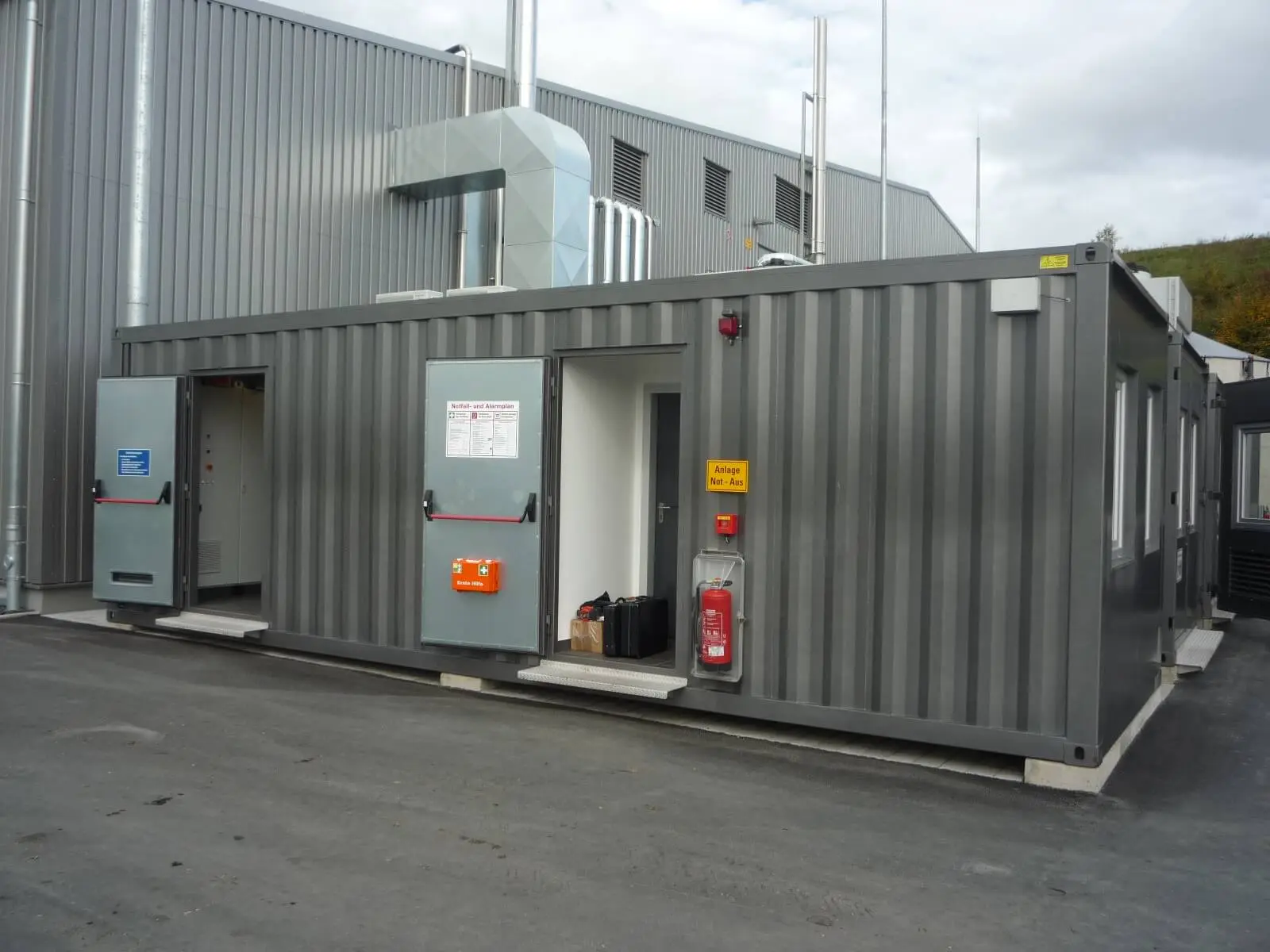 Notstrom-Ersatzanlagen-Container mit Technikraum, Not-Aus-Schalter, Feuerlöscher und geöffneten Wartungstüren an einem Industriegebäude.