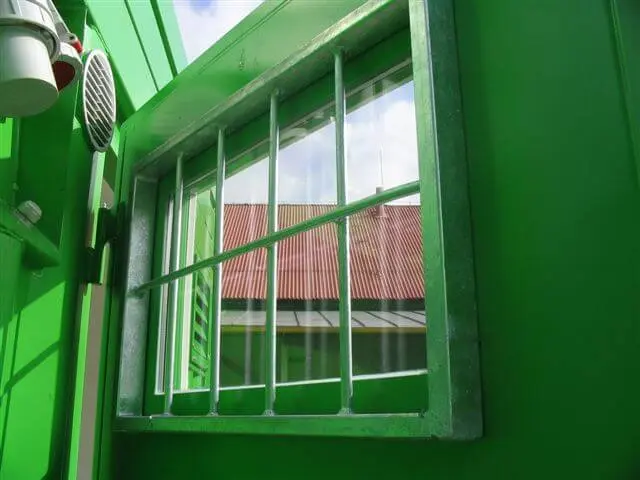 Außenansicht eines grünen Containers mit einem vergitterten Fenster zur Einbruchsicherung, durch das sich ein rotes Ziegeldach spiegelt.